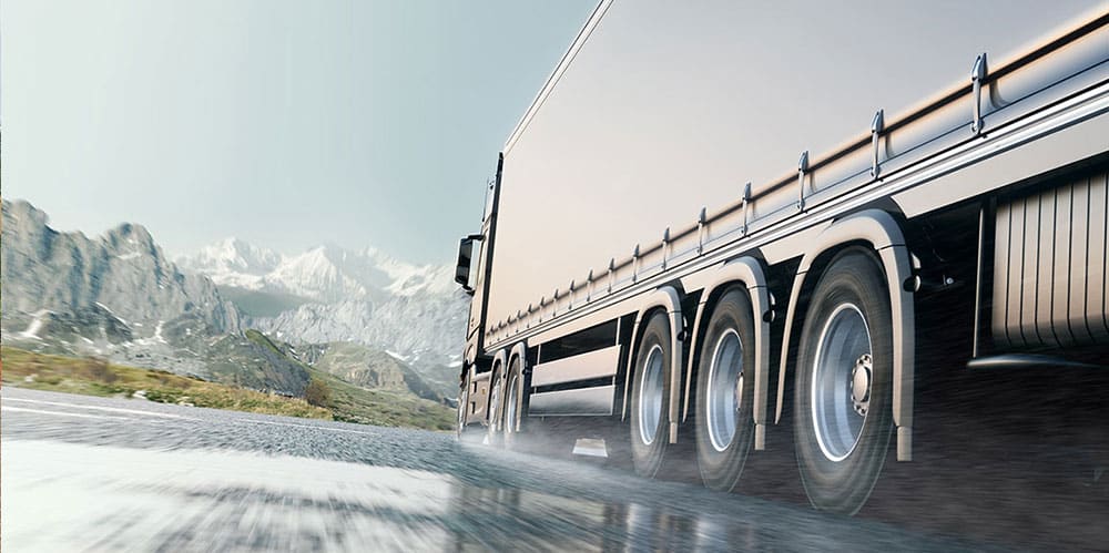 Transport routier : camion circulant dans les montagnes Camion de transport circulant à vive allure sur une route mouillée avec des montagnes enneigées en arrière-plan.